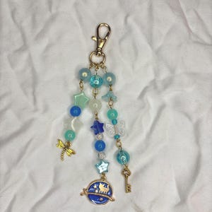 May include: A beaded keychain with a gold clasp. The keychain features blue, teal, and clear beads, along with star, butterfly, and key charms. A blue planet charm with cat silhouettes is also included.