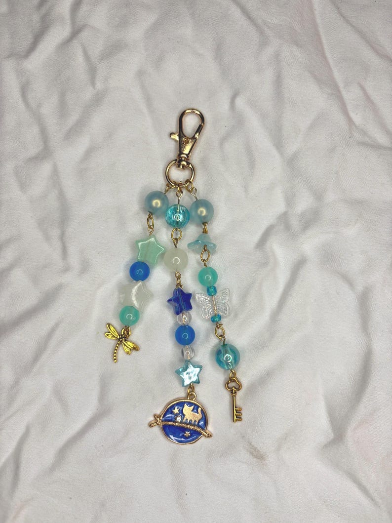 May include: A beaded keychain with a gold clasp. The keychain features blue, teal, and clear beads, along with star, butterfly, and key charms. A blue planet charm with cat silhouettes is also included.