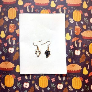 May include: A pair of gold-coloured metal earrings. One earring features a white owl design, and the other has a black cat design. Both earrings have a hook closure. The earrings are displayed on a white surface against a patterned background with pumpkins and autumn foliage.