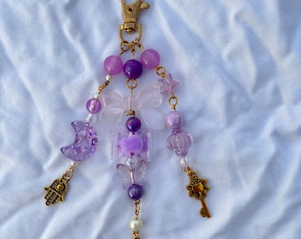 May include: A beaded keychain with a gold clasp and charms. The charms include a purple crescent moon, a Hamsa hand, a key, and a star. The beads are purple and clear, with a butterfly and flower-shaped charm.