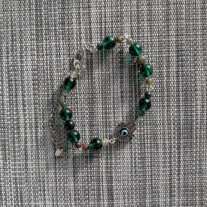 May include: A beaded bracelet featuring dark green and clear beads, with a silver-toned charm of a hand with an eye in the center. The bracelet is on a gray and white textured surface.