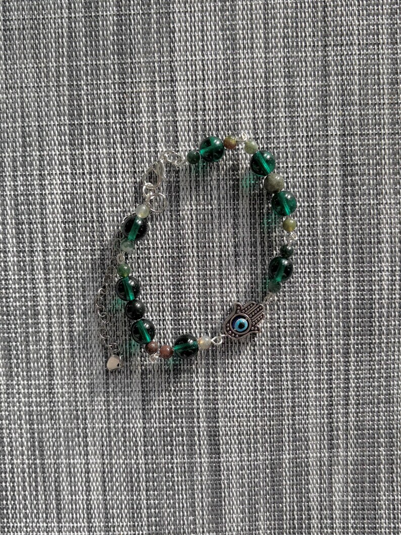 May include: A beaded bracelet featuring dark green and clear beads, with a silver-toned charm of a hand with an eye in the center. The bracelet is on a gray and white textured surface.