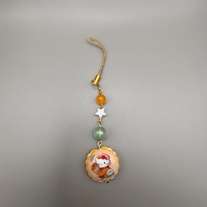 May include: A charm with a gold-colored cord and a circular pendant featuring a cartoon cat wearing a red bow. The charm includes orange, green, and white beads. The pendant has a scalloped edge and a yellow and orange background.