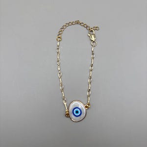 May include: Gold-toned chain bracelet featuring a white and blue evil eye charm. The charm has a white background with a blue eye design in the center. The bracelet has a lobster clasp closure.