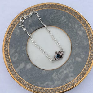 May include: A silver-toned chain bracelet with a Hamsa hand charm, featuring a blue eye design. The bracelet is displayed on a gray and white plate with a gold rim. The Hamsa hand is a symbol of protection.
