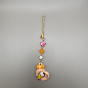 May include: A charm featuring a Hello Kitty design. The charm has a gold-colored string and is adorned with a star-shaped bead, orange and white beads, and a heart-shaped pendant with a bow. The pendant has a Hello Kitty graphic.