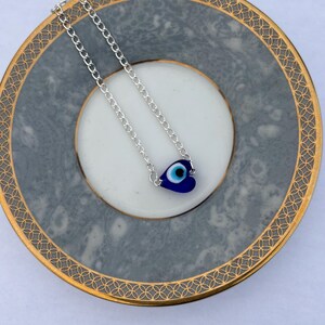 May include: A silver chain necklace with a heart-shaped blue evil eye pendant. The pendant features a white and blue eye design. The necklace is displayed on a gray and gold-rimmed plate.