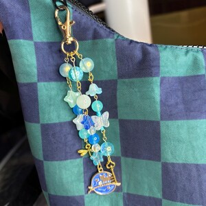 May include: A celestial-themed keychain with a gold clasp, featuring blue and clear beads, star and butterfly charms, and a blue planet pendant with a key. The keychain is attached to a blue and green checkered bag.