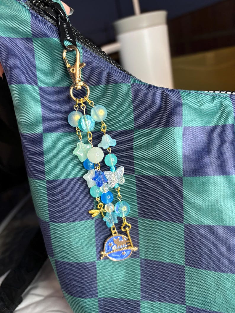 May include: A celestial-themed keychain with a gold clasp, featuring blue and clear beads, star and butterfly charms, and a blue planet pendant with a key. The keychain is attached to a blue and green checkered bag.