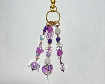 May include: A purple and gold beaded keychain with various charms. The charms include a candy, a star, a butterfly, a flower, a heart-shaped lollipop, and various beads. The keychain has a gold clasp and ring.