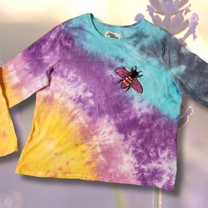 May include: A long-sleeved tie-dye shirt in shades of yellow, purple, and blue. A pink and black bee patch is on the chest. The shirt has a crew neck and a relaxed fit. The background has a bee and lavender.