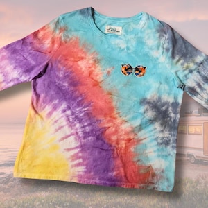 May include: A long-sleeve tie-dye shirt in a vibrant mix of colors, including yellow, purple, red, and blue. The shirt features an embroidered design of sunglasses on the chest. The background shows a sunset and a camper van.