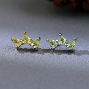 May include: Two pairs of crown-shaped stud earrings. One pair is gold-toned with light green gemstones, and the other is silver-toned with similar green gemstones. The earrings are set against a gray background with a blurred pinecone.