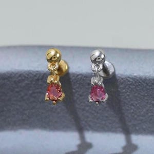 May include: Two stud earrings, one gold-toned and one silver-toned, each featuring a small, dangling, teardrop-shaped pink gemstone. The earrings are displayed on a gray surface, with shadows cast behind them.