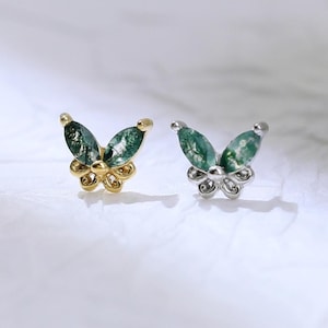 May include: Two butterfly-shaped earrings. One is gold-toned with clear accents, the other is silver-toned with clear accents. Both have green, marbled-effect wings. The earrings are displayed on a white surface.