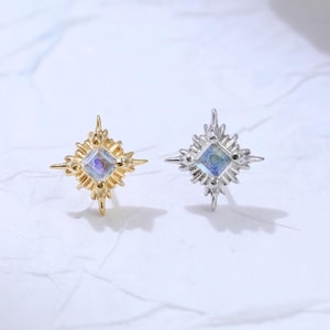 May include: A pair of stud earrings, one gold-toned and one silver-toned, each featuring a central square stone with a starburst design. The stones have a subtle iridescent quality, reflecting various colors.