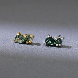 May include: Two pairs of stud earrings with green gemstones. One pair has a gold setting, and the other has a silver setting. The earrings are displayed on a gray surface. The gemstones have a speckled appearance.