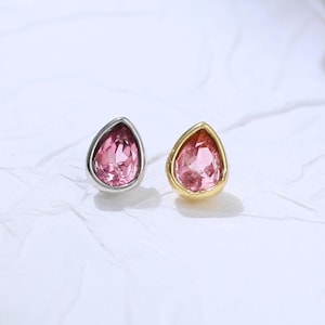 May include: Two teardrop-shaped earrings with pink gemstones. One earring has a silver setting, and the other has a gold setting. The earrings are displayed on a white, textured surface.