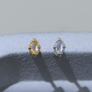 May include: Two small stud earrings, one in gold and one in silver, each featuring a marquise-shaped moonstone set within a beaded metal frame. The earrings are positioned side-by-side, displaying a delicate design with a central iridescent stone.