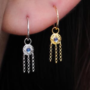 Pode incluir: Close-up de dois brincos, um prateado e outro dourado, cada um com um pequeno centro de pedra preciosa azul e detalhes de corrente pendurada. Os brincos são usados numa orelha, mostrando o seu design e detalhes. Um brinco de argola dourado também é visível.