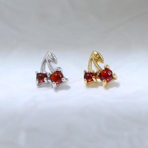 Puede incluir: Dos pendientes en forma de cereza, uno en metal plateado y otro en metal dorado. Cada pendiente presenta dos pequeñas piedras redondas de color rojo oscuro como cerezas, con finos tallos metálicos que se extienden hacia arriba.