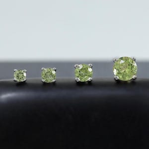 May include: Four peridot stud earrings of varying sizes are displayed on a black, rounded surface. The earrings have a light green hue and are set in silver. The earrings are arranged in a row, showcasing the different sizes available.