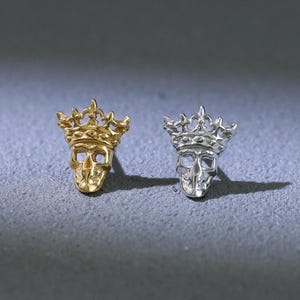 May include: A pair of skull-shaped stud earrings, one gold and one silver, each with a crown. The earrings are presented on a grey surface, highlighting their detailed design and contrasting colours.