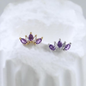 May include: Two stud earrings with a crown design. One earring is gold-toned with purple and white gemstones, and the other is silver-toned with purple and white gemstones. The earrings are displayed on a white, textured surface.