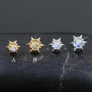 May include: Four stud earrings with a sunburst design, each featuring a central gemstone. Two are gold-toned, and two are silver-toned. The gemstones vary in color, with options labeled A and B. The earrings are displayed on a black, textured surface.