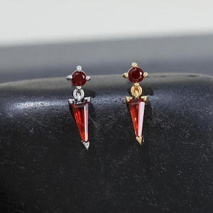 May include: A pair of stud earrings featuring red, triangular gemstone pendants. One earring has a silver setting, the other a gold setting. The earrings are displayed on a black, rounded stone against a neutral background.