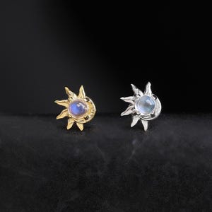 以下が含まれることがあります： 太陽の形をしたスタッドピアス2点。1点はゴールドトーンの金属製、もう1点はシルバートーンの金属製です。それぞれのピアスの中央には丸い虹色のムーンストーンのカボションがあしらわれ、周囲を放射状の太陽光線が囲む天体のようなデザインです。