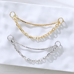 May include: Two chain dangles, one gold-toned and one silver-toned, are displayed on a white surface. Each dangle features two chains connected by small rings. The chains have different textures, adding visual interest to the jewelry.