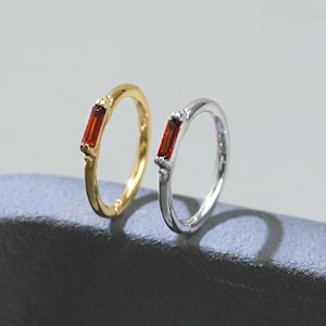 Op de afbeelding: Twee ringen, één goudkleurig en één zilverkleurig, elk met een rechthoekige, rode edelsteen. De gouden ring is links, de zilveren rechts. De ringen staan tegen een grijze achtergrond.