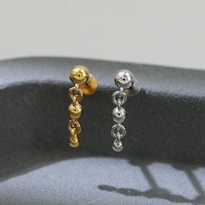 May include: Two stud earrings, one gold-toned and one silver-toned, each featuring a dangling chain of small, round beads. The earrings are displayed on a dark gray surface, with a soft, neutral background.