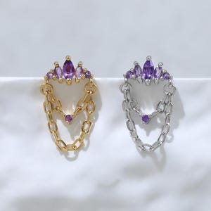 Peut inclure: Deux boucles d'oreilles en forme de couronne, l'une dorée et l'autre argentée, ornées de pierres violettes taille marquise sur le dessus et de deux chaînes pendantes reliées par une petite pierre violette au centre de la boucle inférieure.