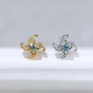Op de afbeelding: Twee oorknopjes met een wervelend, sierlijk metalen ontwerp met een ronde blauwe edelsteen in het midden. De ene oorbel is uitgevoerd in goudkleurig metaal, de andere in zilverkleurig metaal, beide voorzien van gedetailleerde filigraan patronen.