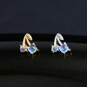 Peut inclure: Une paire de boucles d'oreilles en forme de cerise. L'une est dorée, l'autre argentée. Chaque boucle d'oreille comporte deux pierres précieuses rondes bleu clair. Les boucles d'oreilles sont sur une surface noire brillante.