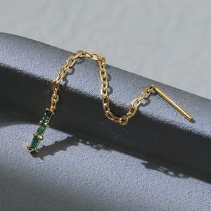 Może przedstawiać: Delikatny, złoty kolczyk z łańcuszkiem i pionowym paskiem z małych, zielonych kamieni szlachetnych. Kolczyk jest prezentowany na ciemnoszarej powierzchni, podkreślając jego elegancki design i kunszt wykonania.