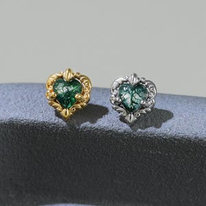 May include: Two heart-shaped stud earrings. One is gold-toned with a green center, the other is silver-toned with a green center. Both earrings have a decorative border. The earrings are displayed on a textured, gray surface.