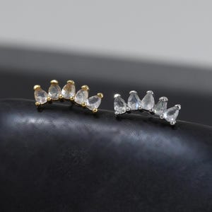 May include: Two curved stud earrings, one in gold-tone metal and one in silver-tone metal, each featuring five clear, pear-shaped stones arranged in a fan-like arc. The earrings are positioned side-by-side, showcasing their metallic finish.