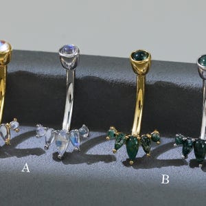 Pode incluir: Quatro piercings no umbigo com barras curvas e detalhes decorativos de pedras preciosas. Dois têm barras douradas e dois têm barras prateadas. As pedras preciosas incluem pedras da lua e pedras de cor esmeralda.