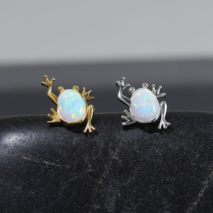 Pode incluir: Dois brincos em forma de sapo, um em metal dourado e outro em metal prateado. Cada brinco apresenta uma pedra de opala branca iridescente de formato oval no centro como o corpo do sapo, com pernas e cabeça detalhadas.