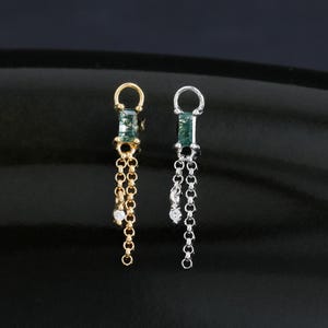 May include: Two dangle earrings, one gold and one silver, each featuring a small green gemstone and a delicate chain. The gold earring has a small white gem at the end of the chain. The earrings are displayed on a black surface.