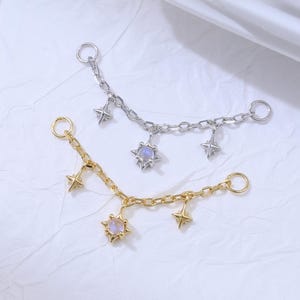 May include: Two delicate chain accessories, one gold-toned and one silver-toned, each adorned with star-shaped charms and a central, iridescent gem. The chains are designed to be worn as body jewelry or as an embellishment.