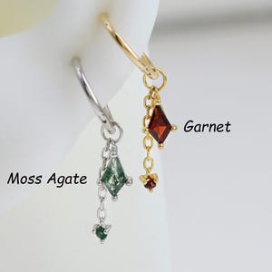May include: Two earrings, one gold and one silver, are displayed. The gold earring features a red, diamond-shaped stone labeled "Garnet." The silver earring has a green, diamond-shaped stone labeled "Moss Agate." Both earrings dangle from a hoop.