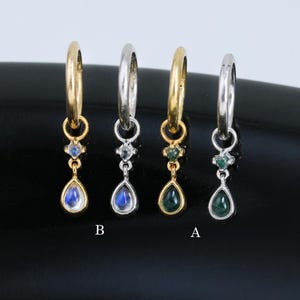 May include: Four pairs of hoop earrings with gemstone pendants. Two pairs have gold hoops, and two have silver hoops. The gemstones are blue and green teardrop shapes, with smaller accent stones above. The earrings are displayed on a black surface.