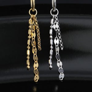 May include: Two hoop earrings, one gold-toned and one silver-toned, each featuring a trio of dangling chains. The chains consist of small, flat, oval-shaped links that hang from a circular hoop, creating a layered, delicate appearance.