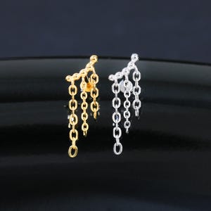 May include: A pair of earrings, one gold and one silver, each featuring a cluster of dangling chain links. The earrings are displayed on a reflective black surface, creating a contrast between the jewelry and the background.