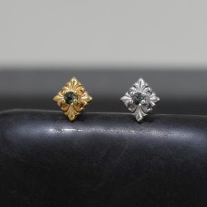 Peut inclure: Deux boucles d'oreilles à tige en forme d'étoile à quatre branches ou de croix avec un travail de métal orné de style filigrane. Une boucle est dorée et l'autre argentée, chacune sertie d'une petite pierre précieuse ronde et sombre au centre.