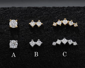 May include: A collection of six stud earrings arranged in two rows on a black display card. The top row features gold-tone settings, while the bottom row features silver-tone settings. Designs include single stones, double stones, and curved multi-stone clusters.